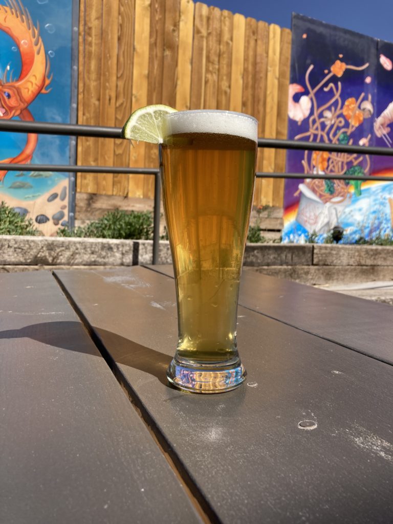 photo of tall glass of light colored beer, La Llagerona, with a lime wedge on a picnic table